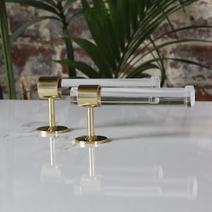 Two brass-colored metal fixtures with clear glass tubes attached, placed on a white surface against a brick wall background.