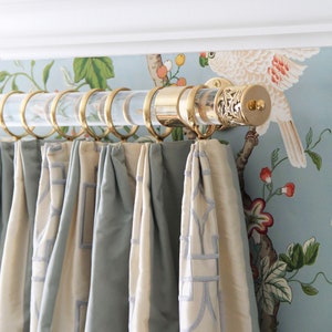 1.5 inch round lucite rod with brass hardware, wall mounted on a light blue patterned wall, holding a linen drape with braided trim.