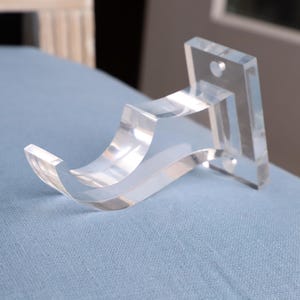 May include: A clear acrylic curtain rod bracket with a curved arm and a square base. The bracket is designed to hold a curtain rod.