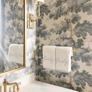 Vignette of a traditional looking powder room with grey tree patterned wallpaper and gold accessories around the sink. Two white monogrammed towels are draped over a clear lucite and satin brass towel rod.