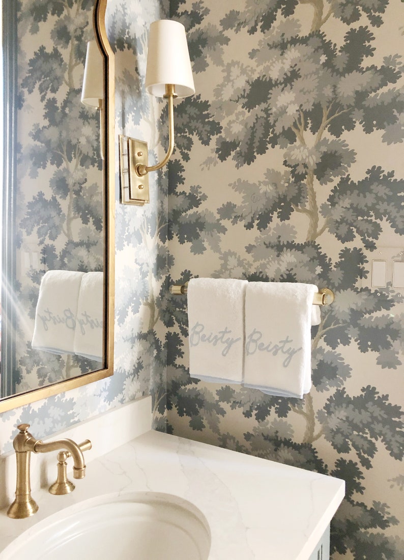 Vignette of a traditional looking powder room with grey tree patterned wallpaper and gold accessories around the sink. Two white monogrammed towels are draped over a clear lucite and satin brass towel rod.