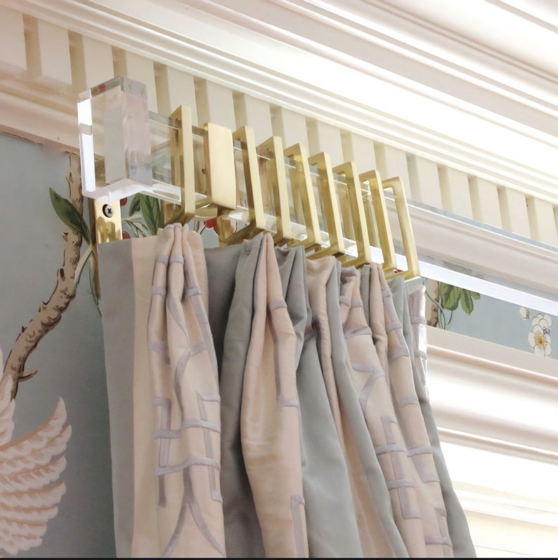 Rectangular lucite curtain rod with gold brass rings and clear acrylic rectangle end caps, holding up neutral draperies in front of a patterned wallpaper wall.