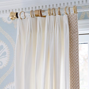 A round lucite curtain rod with gold curtain rings, holding up off white drapes, featuring imperial style end cap in brass.