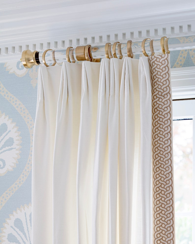 A round lucite curtain rod with gold curtain rings, holding up off white drapes, featuring imperial style end cap in brass.