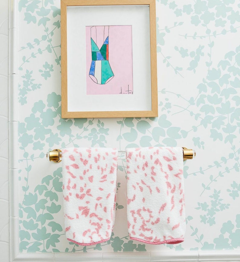 A clear lucite and polished brass towel bar holds two white and pink hand towels. The towel bar is wall mounted under artwork on a light green foliage style wallpaper.