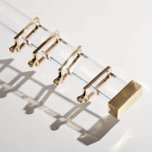 Rectangular lucite curtain rod with gold brass rectangular rings, holding up a grey draperies in front of a blank white wall.