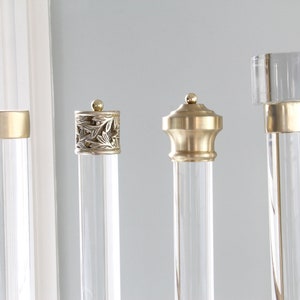 May include: Four different finials for curtain rods. The finials are made of clear acrylic and have gold metal accents. The finials are all different styles, including a simple round finial, a finial with a decorative design, a finial with a square top, and a finial with a round top.