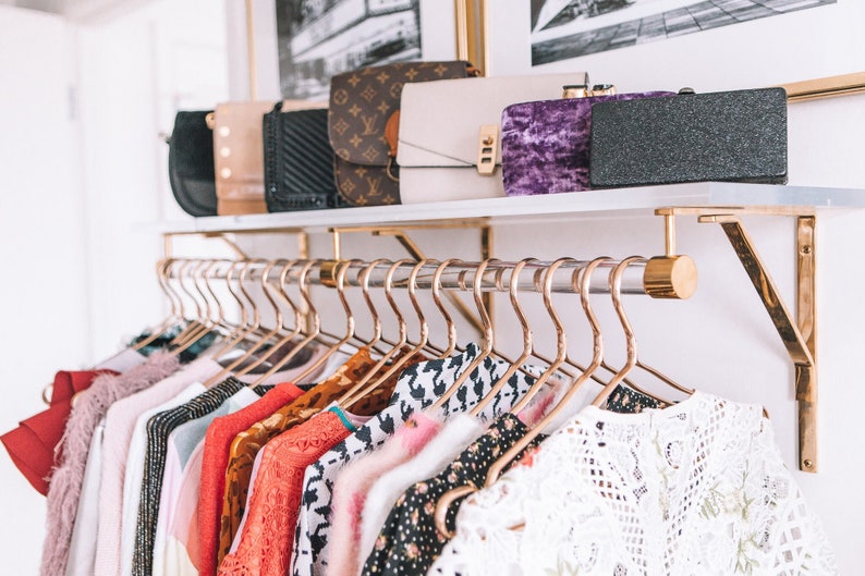 Lucite shelf with brass brackets, holding a clear acrylic rod with matching hardware, used to hang clothes. A variety of clutches and purses are displayed on the top of the shelf.