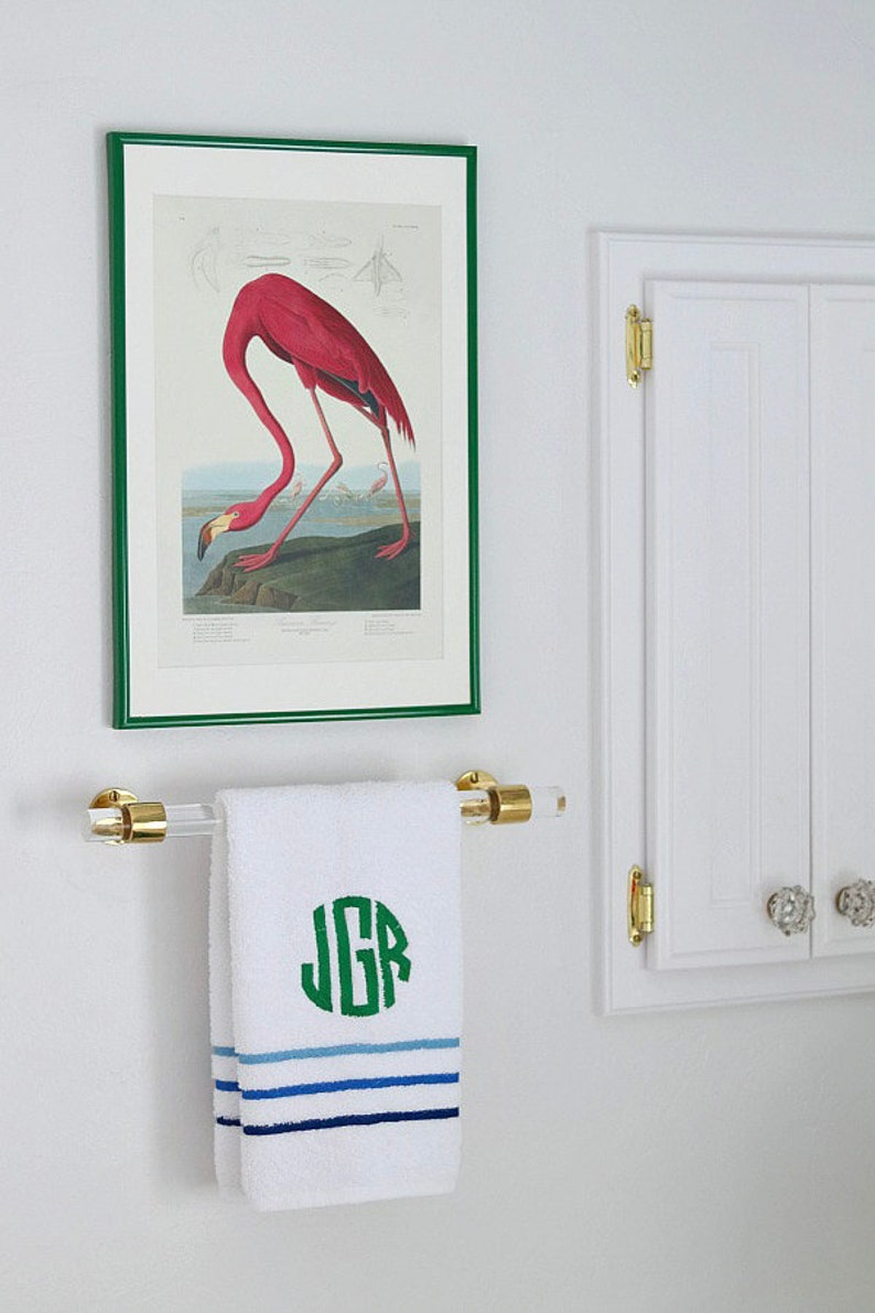 A clear lucite acrylic and polished brass towel rod is installed on the wall of a bathroom, holding a modern monogrammed hand towel. Above the towel is a piece of artwork featuring a flamingo, and next to the towel bar is a medicine cabinet.