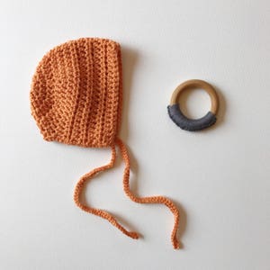 May include: An orange crochet baby bonnet with tie strings and a wooden teething ring with a gray fabric accent. The bonnet is made of a textured yarn and is a warm, earthy color. The teething ring is a natural wood color.
