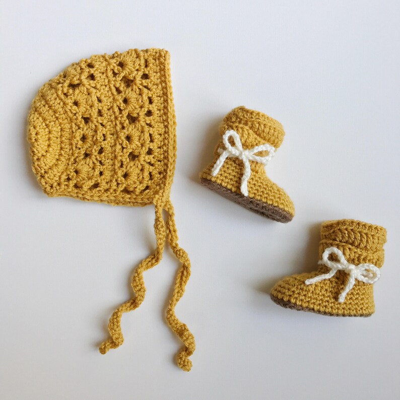 May include: A set of crocheted baby booties and a bonnet in a mustard yellow color. The booties have white bows and the bonnet has a long tie.