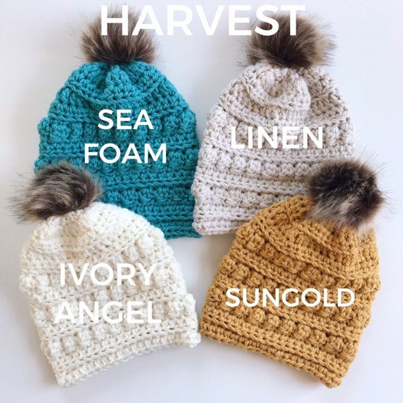 May include: Four handmade crochet beanies in various colors: sea foam, linen, ivory angel, and sungold. Each hat features a textured stitch pattern and a faux fur pom-pom on top. The word "HARVEST" is at the top of the image.