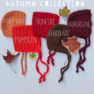 May include: An assortment of crocheted baby bonnets in autumn colors: Honey Gold, Pumpkin, Bonfire, Chocolate, and Aubergine. Each bonnet has a tie closure and is displayed with fall leaves. The text "AUTUMN COLLECTION" is at the top.