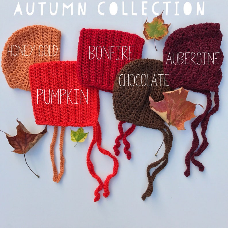 May include: An assortment of crocheted baby bonnets in autumn colors: Honey Gold, Pumpkin, Bonfire, Chocolate, and Aubergine. Each bonnet has a tie closure and is displayed with fall leaves. The text "AUTUMN COLLECTION" is at the top.