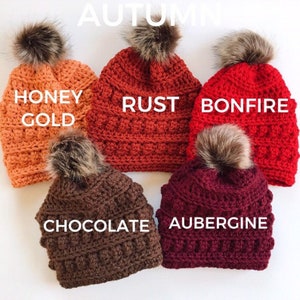 May include: Five crocheted beanies in autumn colors: Honey Gold, Rust, Bonfire, Chocolate, and Aubergine. Each hat has a faux fur pom-pom on top. The word "AUTUMN" is at the top of the image.