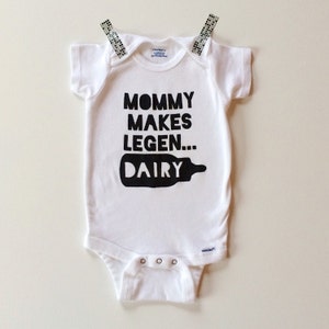 May include: White baby onesie with the text "MOMMY MAKES LEGEN... DAIRY" in black. The onesie is held up by two decorative clips. The onesie has short sleeves and snaps at the bottom. The onesie is made of a soft material.