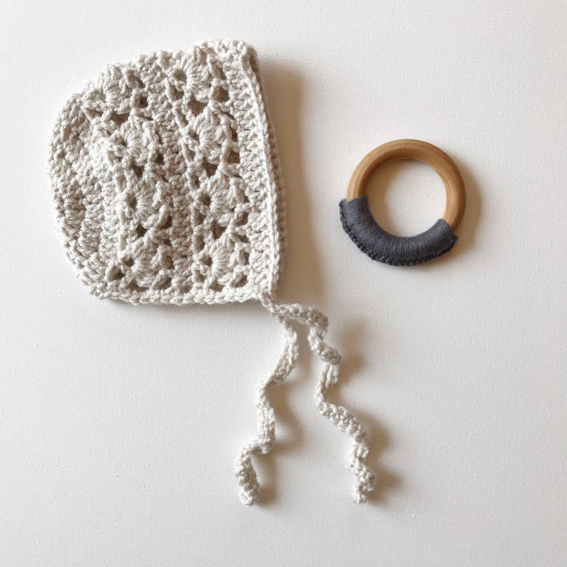 May include: A crocheted baby bonnet with a white crocheted tie. The bonnet is made with a delicate, lacy pattern. A wooden teething ring with a gray fabric band is also pictured.