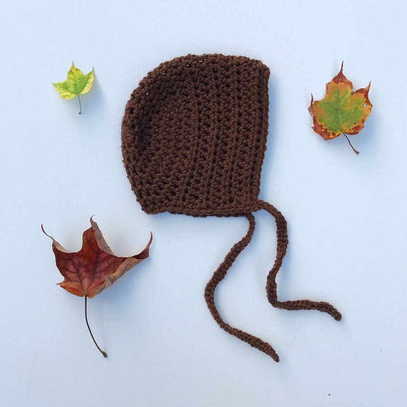 May include: A handmade brown crochet baby bonnet with tie strings, set against a light blue background. The bonnet is surrounded by three colorful autumn leaves, creating a cozy, seasonal aesthetic. The bonnet is a warm, earthy brown color.
