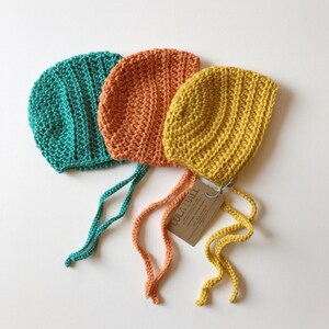 May include: Three handmade crochet bonnets in teal, orange, and yellow. Each bonnet has a rounded crown and long ties. A brown tag is attached to the yellow bonnet. The bonnets are displayed on a white surface.