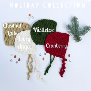 May include: A collection of four crocheted baby bonnets in holiday colors: Chestnut Latte, Ivory Angel, Mistletoe, and Cranberry. Each bonnet is labeled with its color name. The bonnets are displayed on a white surface with festive decorations and a sprig of evergreen.