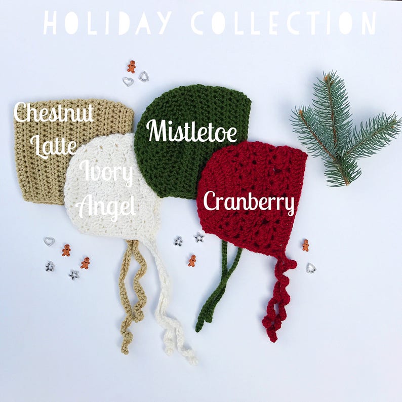 May include: A collection of four crocheted baby bonnets in holiday colors: Chestnut Latte, Ivory Angel, Mistletoe, and Cranberry. Each bonnet is labeled with its color name. The bonnets are displayed on a white surface with festive decorations and a sprig of evergreen.