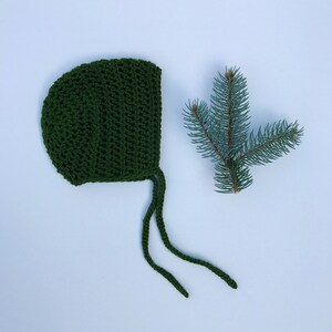 May include: A dark green, crocheted baby bonnet with long ties, next to a sprig of evergreen needles. The bonnet is made of a textured yarn and has a rounded shape. The background is a light blue.