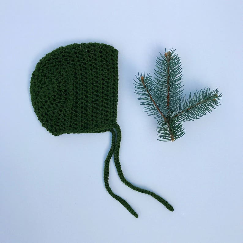 May include: A dark green, crocheted baby bonnet with long ties, next to a sprig of evergreen needles. The bonnet is made of a textured yarn and has a rounded shape. The background is a light blue.