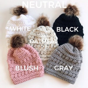May include: Five crocheted beanies with faux fur pom poms in various neutral colors: white, black, oatmeal, blush, and gray. The beanies are arranged in a semi-circle on a white background. The word "NEUTRAL" is written above the beanies.