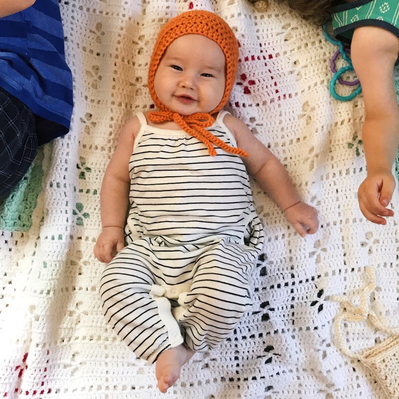 May include: A baby wearing an orange crocheted bonnet with a matching tie, and a black and white striped romper. The baby is lying on a white crocheted blanket.