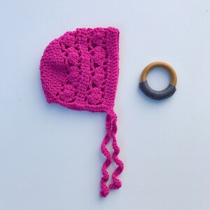 May include: A pink crocheted baby bonnet with a long tie. The bonnet is on a white surface next to a wooden teething ring with a gray fabric covering.