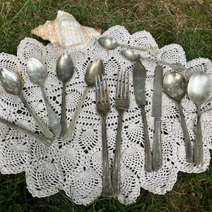 May include: An assortment of vintage silverware, including spoons, forks, and knives, arranged on a white lace doily. A seashell rests in the background. The silverware has a tarnished, antique appearance, suggesting age and use.