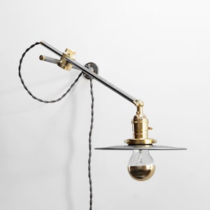 May include: A silver and brass wall-mounted adjustable lamp with a black cord and a round, silver shade. The lamp has a single light bulb.