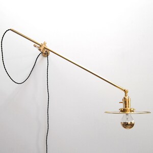 May include: A brass wall-mounted light fixture with a long, adjustable arm and a round, flat shade. The light fixture has a black cord and a simple design.