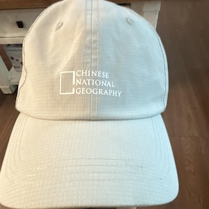 May include: A light beige baseball cap with a grid-like texture throughout the fabric. The front features a white embroidered rectangular logo outline followed by the text CHINESE NATIONAL GEOGRAPHY in white capital letters on the center panel.