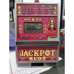 May include: A gray plastic Jackpot Slot machine toy with a digital screen displaying a win. The front features a red panel with yellow text, a payout table, and blue buttons labeled Bet, Spin, and Bell. The base displays the text Jackpot Slot.