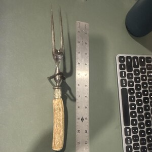 以下が含まれることがあります： 質感のある明るい色のハンドルと装飾的な銀色の襟を備えた、2本歯の金属製カービングフォーク。フォークの横に置かれた金属製の定規は、調理器具の全長が約28センチメートルであることを示しています。