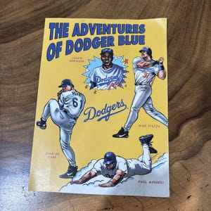 May include: A yellow poster titled The Adventures of Dodger Blue featuring illustrations of baseball players Jackie Robinson, Chan Ho Park, Mike Piazza, and Raul Mondesi in white and blue uniforms with the Dodgers team logo in blue text.