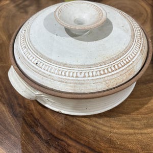 May include: A round, off-white ceramic casserole dish with a matching lid. The lid features a central knob handle and a decorative embossed beaded border. The base has two small side handles and a smooth, light-colored finish with brown accents.
