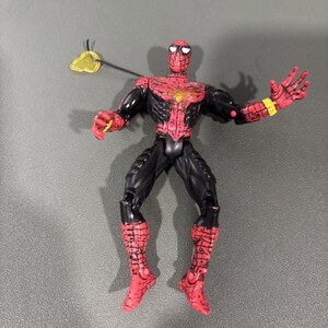 May include: A posable action figure of a superhero character with a red and black suit featuring a spider web pattern. The figure has a gold spider emblem on the chest, yellow wrist bands, and a translucent yellow web accessory attached to the shoulder.