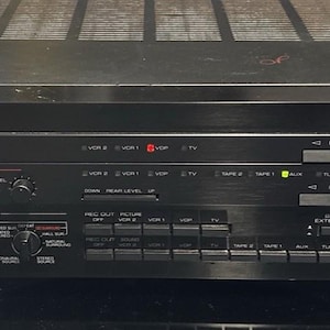 Op de afbeelding: Een zwarte Yamaha Natural Sound stereo-versterker AVC-50 met een grote volumeknop aan de rechterkant, meerdere in- en uitgangsknoppen, een beeldselector en een audio-video-ingangspaneel aan de voorzijde met rode, witte en gele poorten.