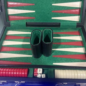 May include: A backgammon set in a black case with a green felt interior featuring red and white triangular points. The set includes two black leather dice cups, a stack of red checkers, a stack of white checkers, and four dice, one marked with the number 2.