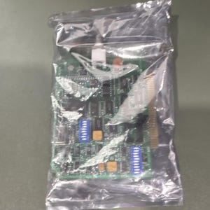May include: A green rectangular circuit board enclosed in a clear plastic bag. The electronic component features multiple black integrated circuits, two blue DIP switches, and a gold-coloured edge connector along the right side of the board.