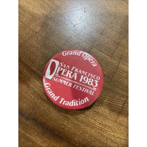 Op de afbeelding: Een ronde rode pinback-button met witte tekst die Grand Opera, San Francisco Opera 1983 Summer Festival en Grand Tradition leest. Het ontwerp bevat een gestileerde letter O en decoratieve lijnen rondom de centrale tekst.