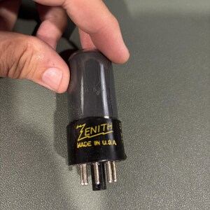 May include: A vintage Zenith vacuum tube with a dark glass envelope and a black base. The base features yellow printed text that reads Zenith and Made in U.S.A. The tube has several metal pins extending from the bottom for electrical connection.