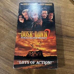 May include: A rectangular VHS tape case for the movie From Dusk Till Dawn 2: Texas Blood Money. The cover features a dark, dramatic sunset landscape with a building, a flock of bats, and a montage of five actors' faces at the top.