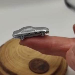 Puede incluir: Un pequeño coche de juguete plateado con cuatro ruedas. El vehículo en miniatura se sostiene entre dos dedos, con una moneda de un centavo y una rodaja de madera en el fondo. El coche tiene un diseño elegante y aerodinámico.