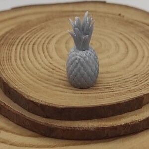 May include: A small, gray pineapple figurine with detailed texture, set on a wooden slice. A penny is in the foreground for size comparison. The pineapple has a textured body and spiky top.