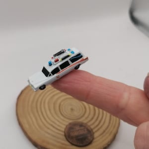 Può includere: Un'auto dei Ghostbusters bianca in miniatura con accenti rossi e blu e una trappola per fantasmi sul tetto. L'auto è appoggiata su un dito.