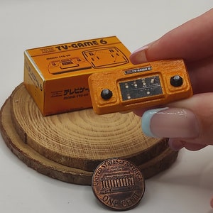 Puede incluir: Una consola de videojuegos de mano en miniatura de color naranja y negro con el texto "TV-GAME 6" en la parte delantera. La consola está sobre una superficie de madera junto a una pequeña caja con el mismo texto.