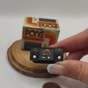 Puede incluir: Mini consola de juegos Atari negra con detalles dorados, sostenida frente a una caja de juego Pong vintage. La consola tiene el logotipo de Atari y detalles de los altavoces. Una moneda está al lado de la consola. La caja de Pong tiene un diseño retro.